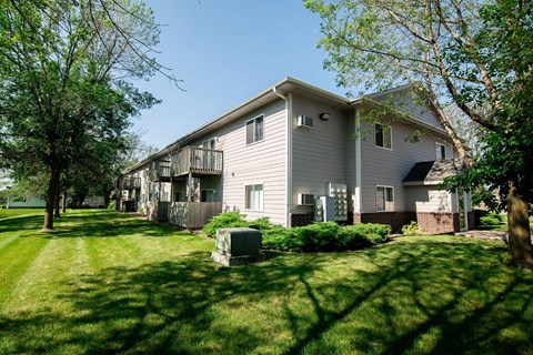 A house with a grey siding and a balcony is surrounded by green grass and trees.