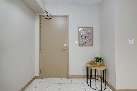 A room with a brown door, a small table with a plant and a picture on the wall.