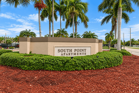 A sign for South Point Apartments is surrounded by a hedge and palm trees.