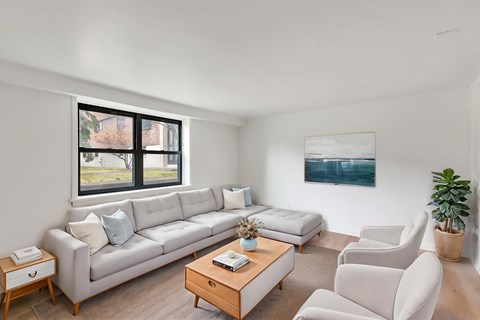 A living room with a large window, a white couch, a coffee table, and a painting on the wall.