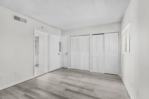 A room with white walls and wood flooring with a door on the left and three closets on the right.