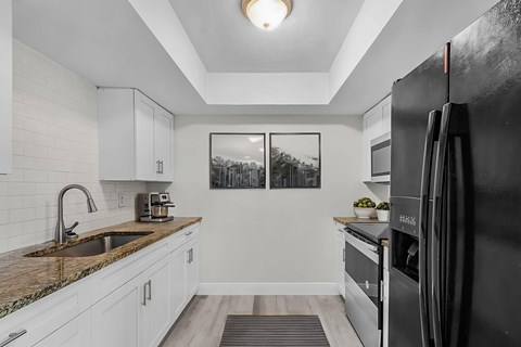 A black refrigerator stands in a kitchen with white cabinets and a granite countertop.