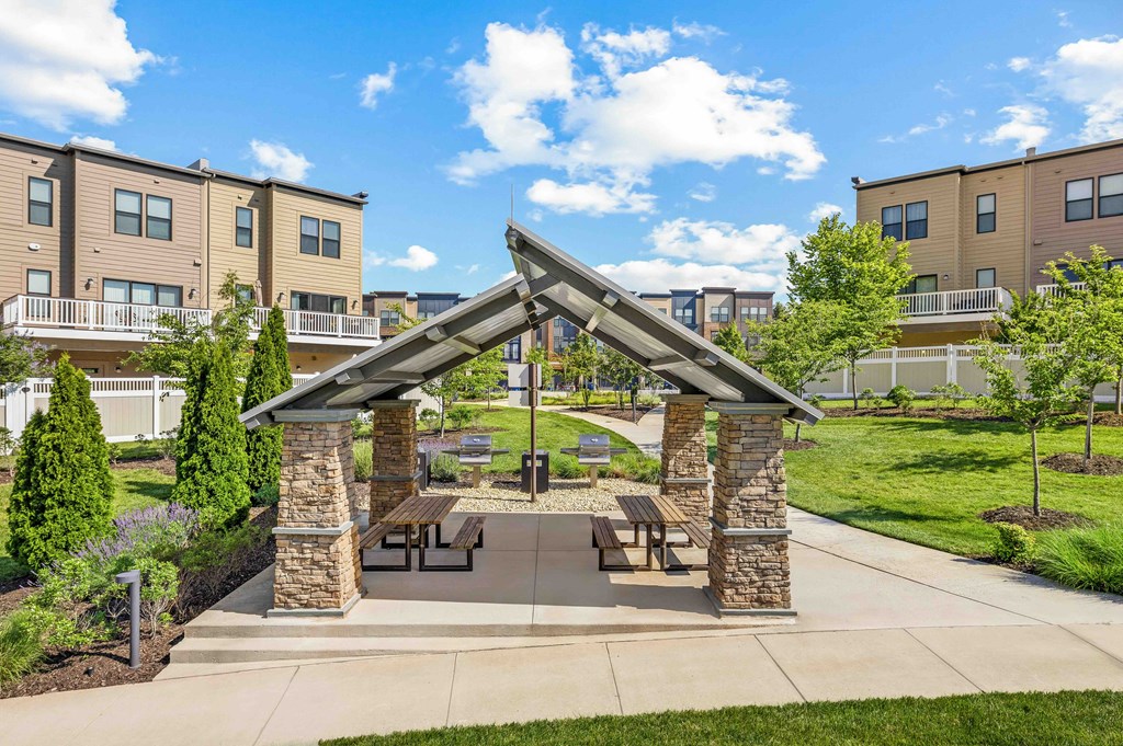 A pavilion with a stone pillar and a roof is surrounded by benches and a lawn.