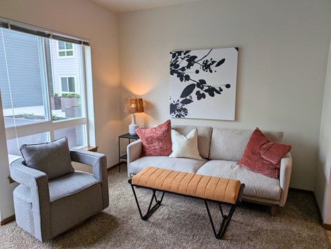 A living room with a grey couch, two grey armchairs, a small table with a lamp, and a large abstract painting on the wall.