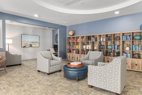 A living room with a grey patterned armchair, a blue ottoman, a wooden bookshelf, and a painting on the wall.
