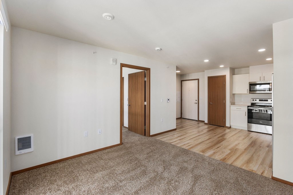 A spacious living room with a carpeted floor and a kitchen area in the background.