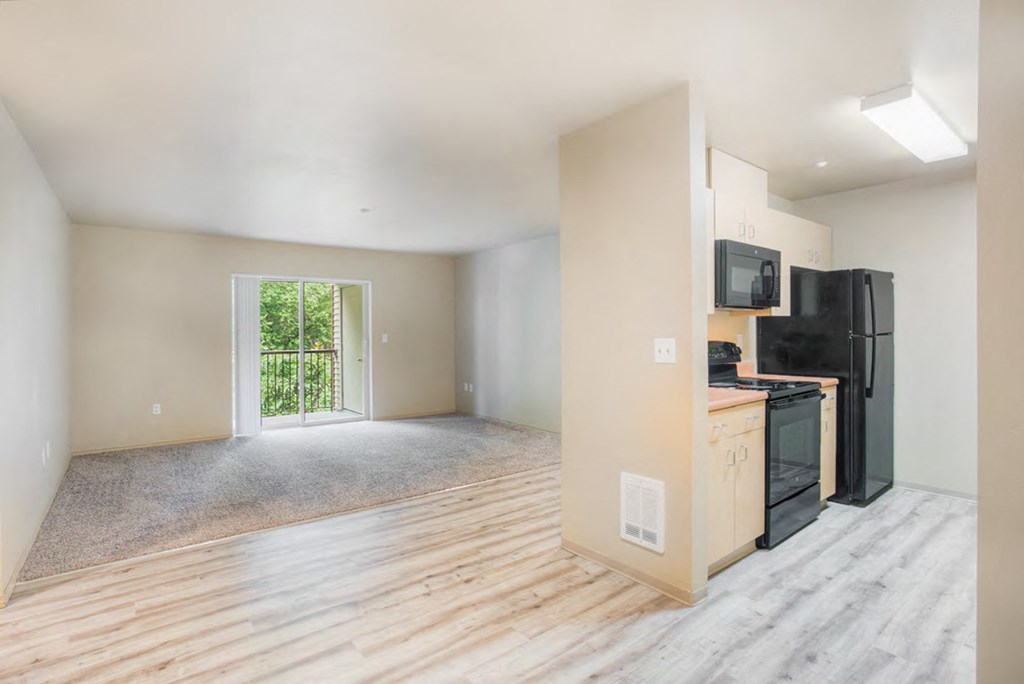 an empty living room with a kitchen and a black refrigerator