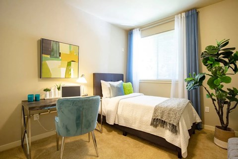 A bedroom with a bed, desk, chair, and potted plant.