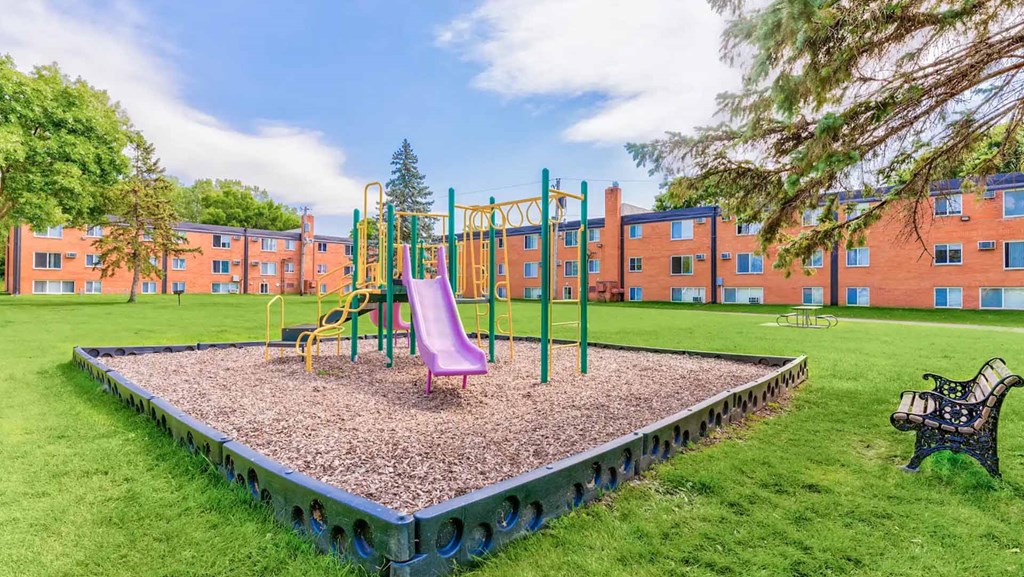 A playground with a slide, swings, and a seesaw in front of a building.