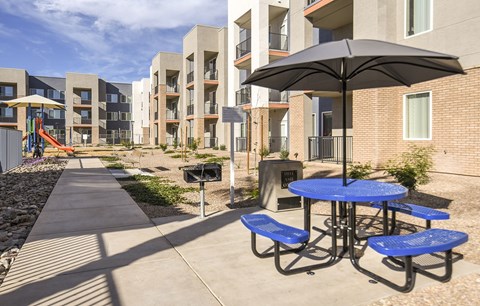 A playground with a slide and a picnic table with blue benches is in front of apartment buildings.