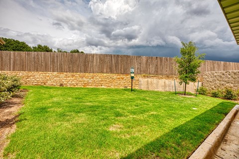 a backyard with a brick wall and a green lawn at Sylvia at South Broadway 55+ Apartments, Joshua, TX 76058
