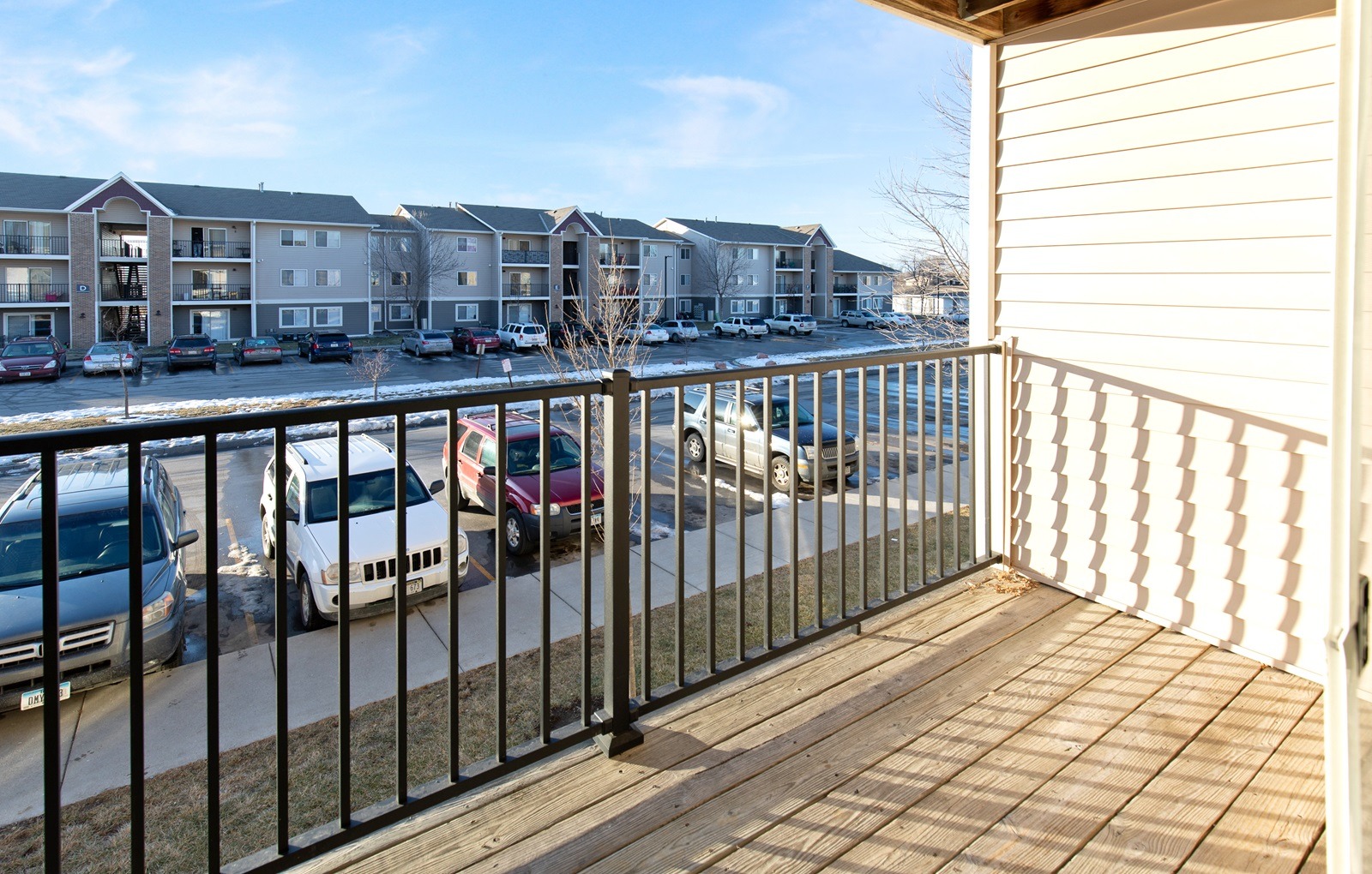 A balcony with a black railing and a view of a parking lot with cars and apartment buildings.