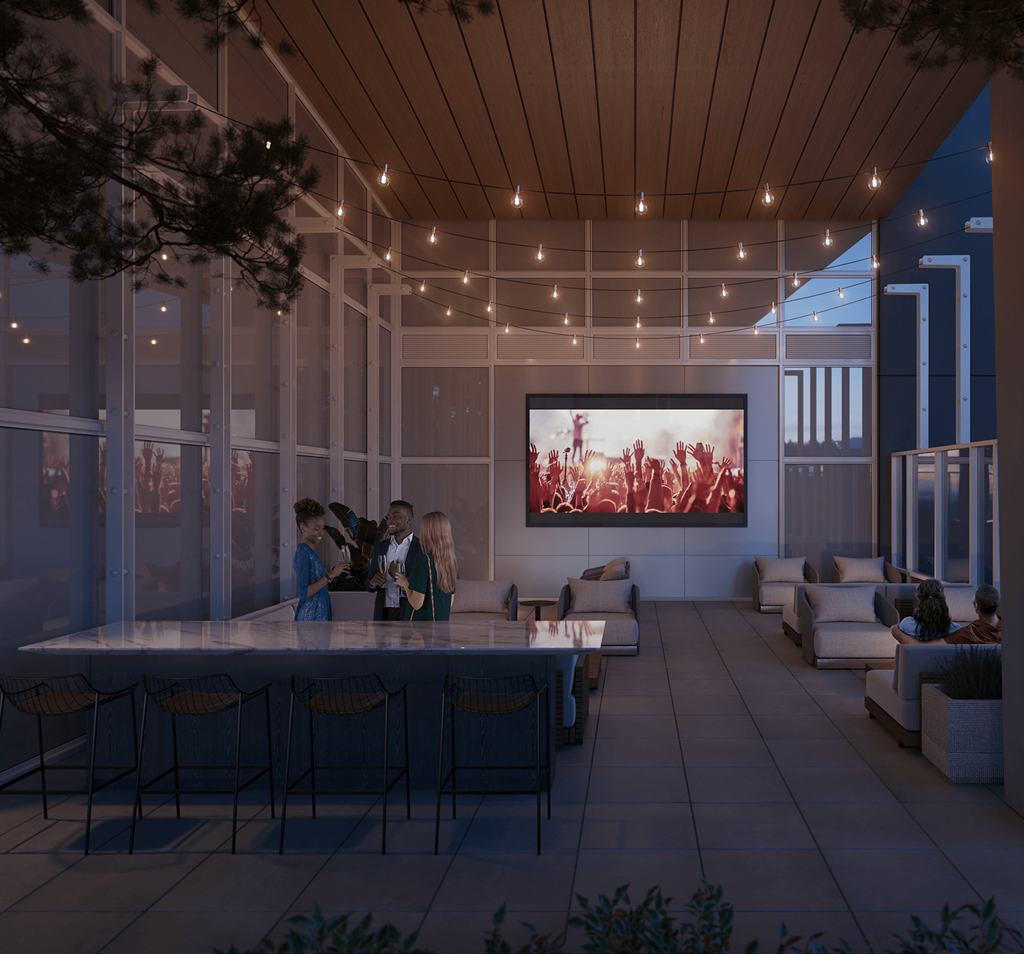 A group of people are gathered around a table in a modern, well-lit room with a large screen displaying a sunset scene.