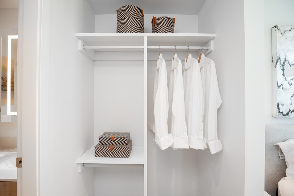 A white closet with clothes hanging and a shelf with boxes and a basket on it.