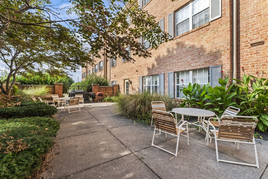 A patio with a table and chairs is surrounded by a brick building and greenery.