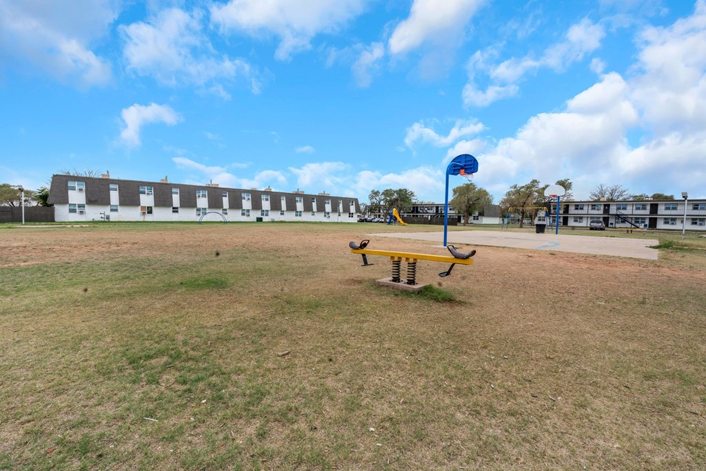 A yellow seesaw in the middle of a grassy field with a blue basketball hoop in the background.