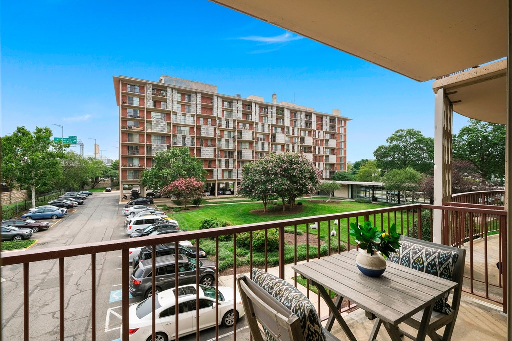 A balcony with a table and chairs overlooks a parking lot and apartment building.