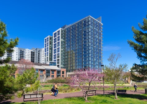 A modern glass building is surrounded by trees and people are walking around.