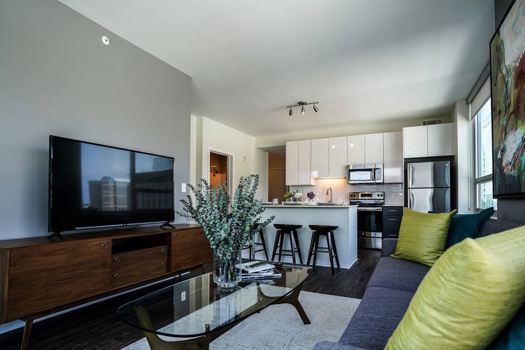 A modern living room with a television, a glass coffee table, and a kitchen in the background at Two Twelve Clayton Apartments, Clayton, MO