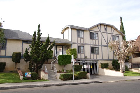 A building with a grey and white facade is surrounded by greenery and a metal gate.