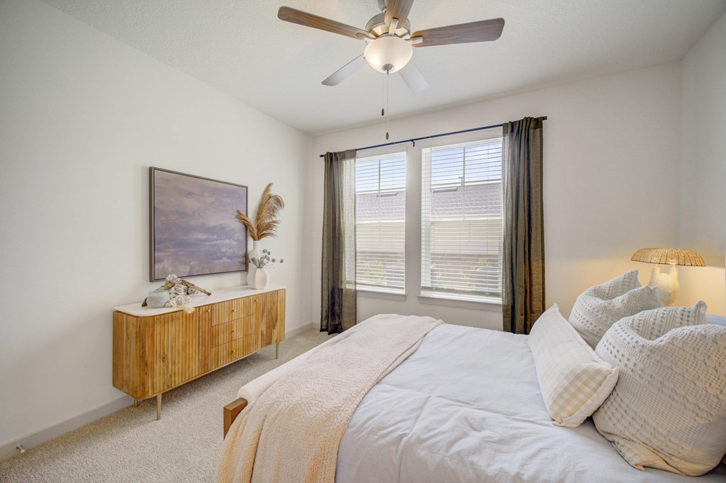 A bedroom with a bed, a ceiling fan, and a wooden cabinet with a painting on top.