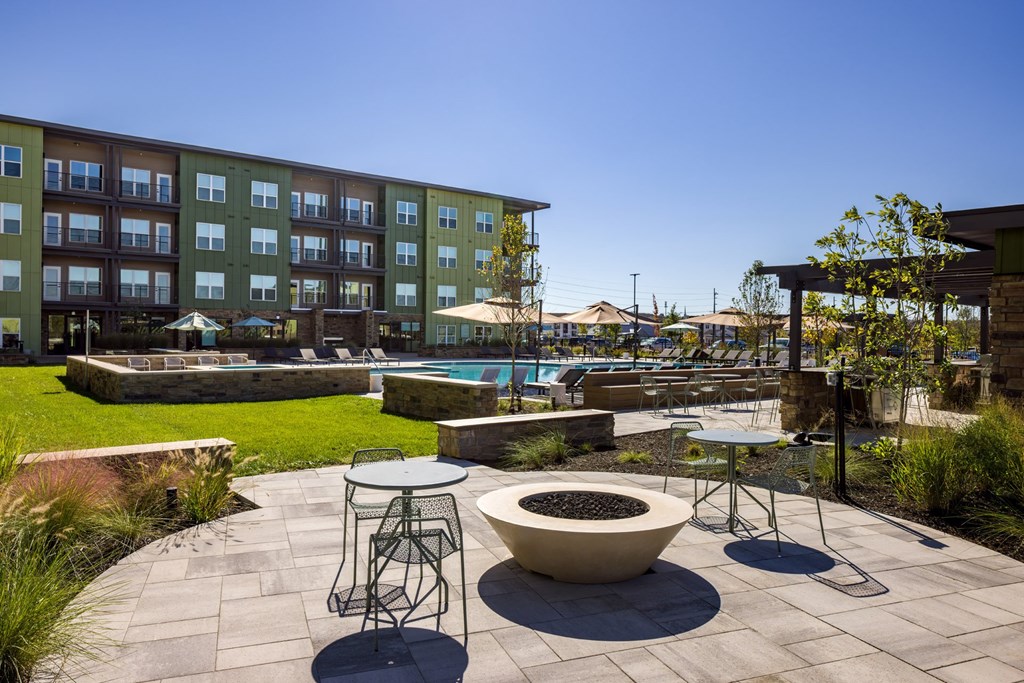 A patio with a fire pit and chairs is surrounded by a grassy area and a building.