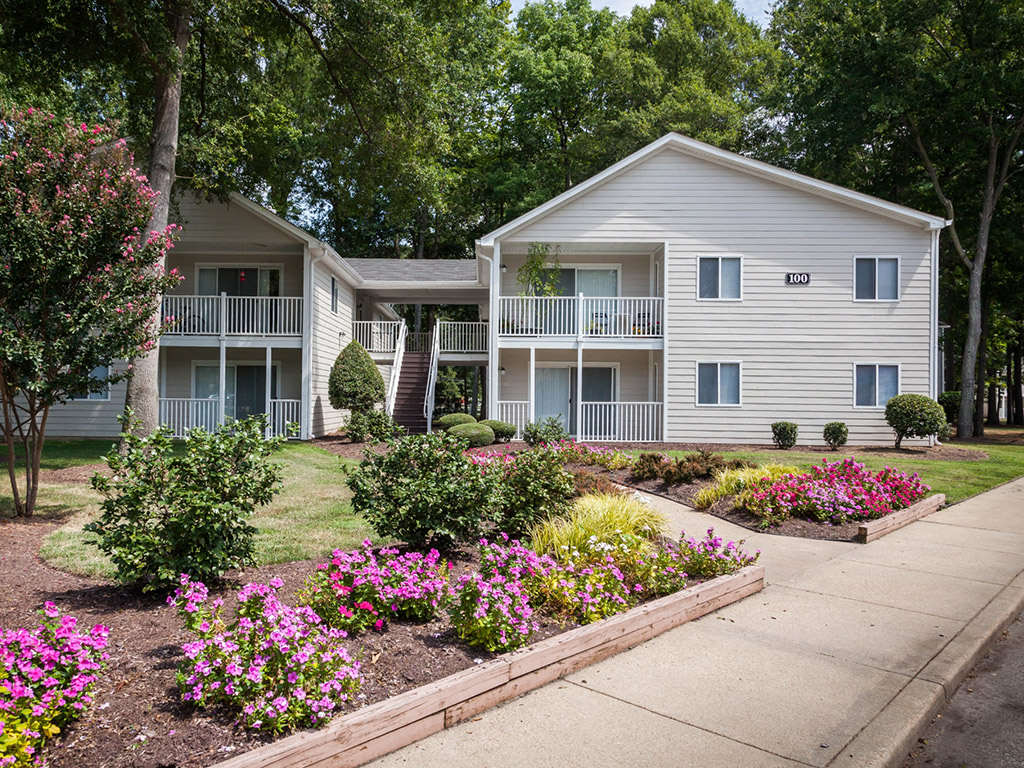 A white two-story apartment building with a balcony and a garden in front.