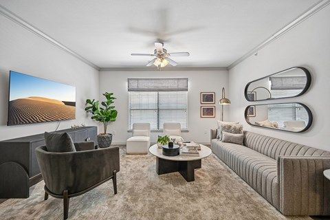 A living room with a grey sofa, a grey armchair, a coffee table, a potted plant, a painting, a mirror, and a ceiling fan.