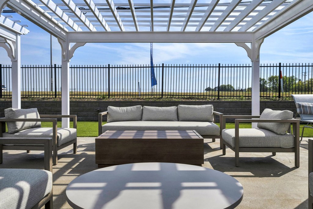 A white pergola with a wooden coffee table and grey couches.