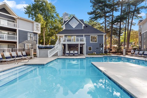 A swimming pool in front of a house with a deck and lounge chairs.