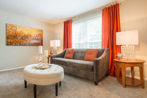 A living room with a grey couch, orange pillows, a round ottoman, and a painting on the wall.