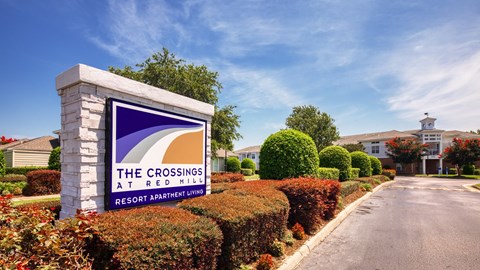 A sign for The Crossings at Red Mill Resort Apartment Living stands in front of a row of bushes.