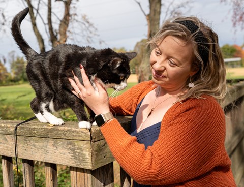 A woman in an orange sweater is petting a black and white cat on a wooden fence.