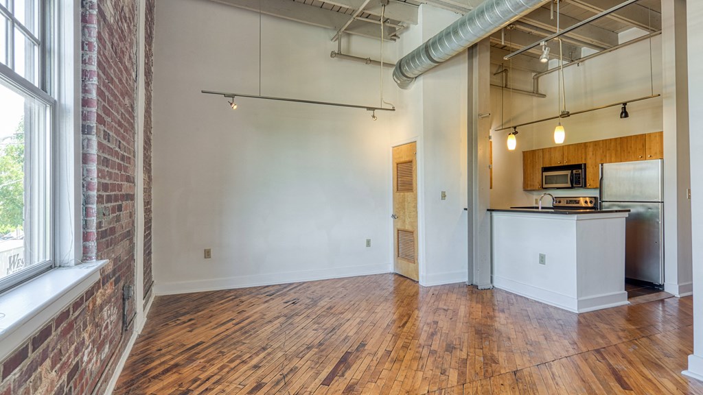 an empty living room with a kitchen and a exposed brick wall