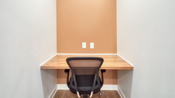 A black chair is in front of a wooden desk with a white wall and two switches on the wall.