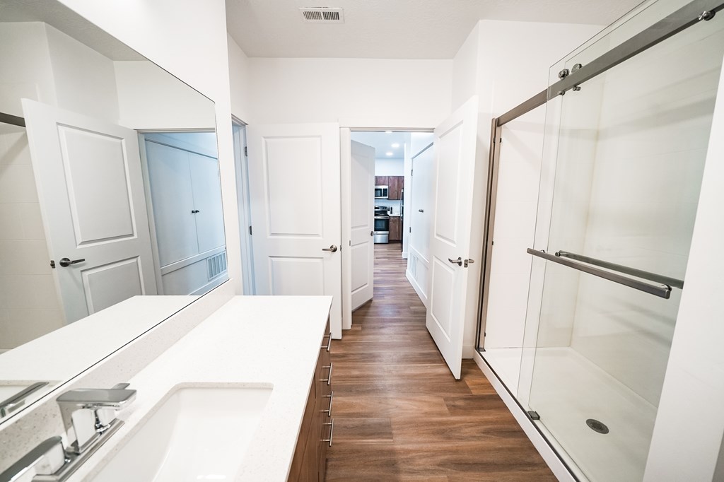 A long white bathroom with a sink and mirror on the left and a walk in shower on the right.
