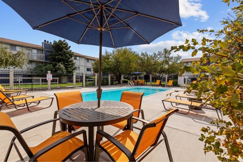 A table with chairs and an umbrella is in the foreground of a pool area.