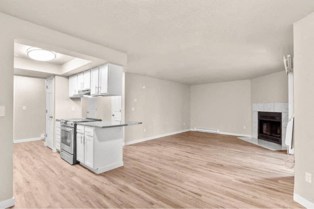 A large, empty living room with a fireplace and a kitchen area with white appliances.