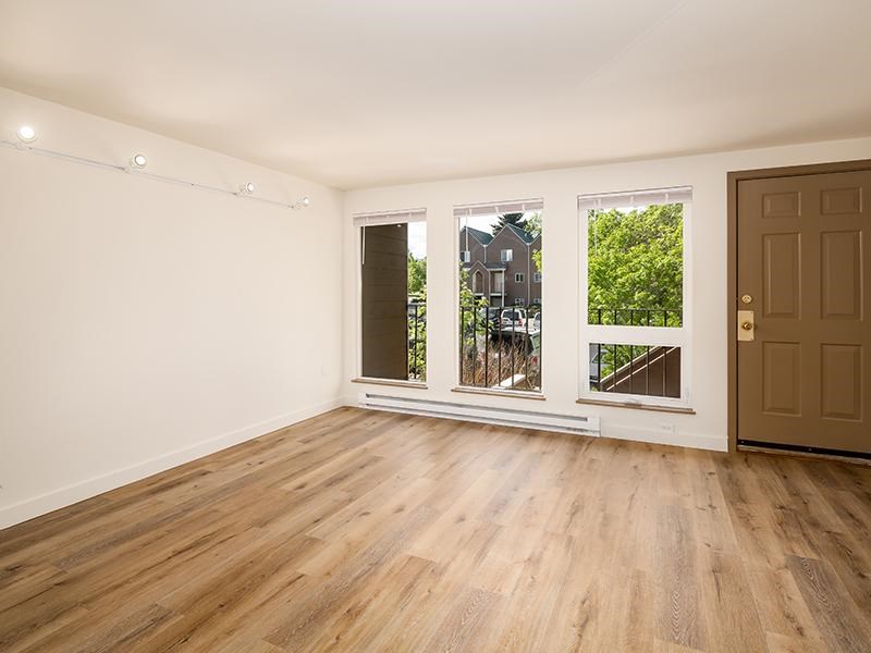A room with wooden floors and white walls, with a brown door and windows showing a view of a house and trees.