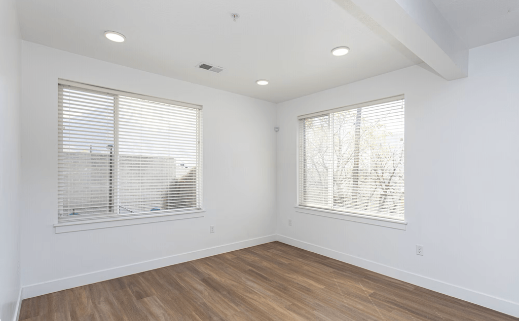 A room with wooden floors and two windows with blinds.
