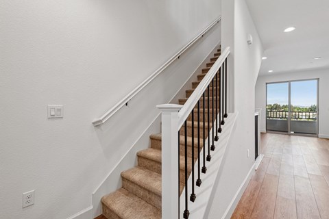 A staircase with a beige carpeted runner and black balusters leads to a bright room with a sliding glass door.