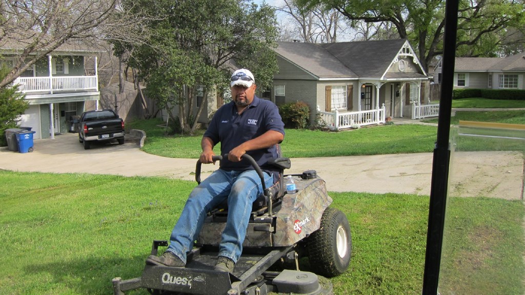 A man in a blue shirt and jeans is riding a Quest lawn mower.
