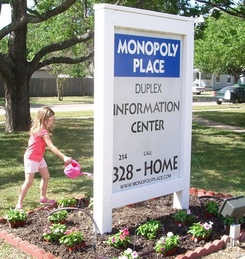 A girl watering flowers in front of a Monopoly Place sign.