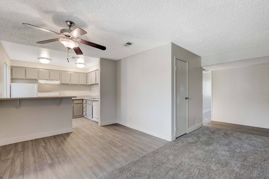 A room with a ceiling fan and a kitchen area in the background.