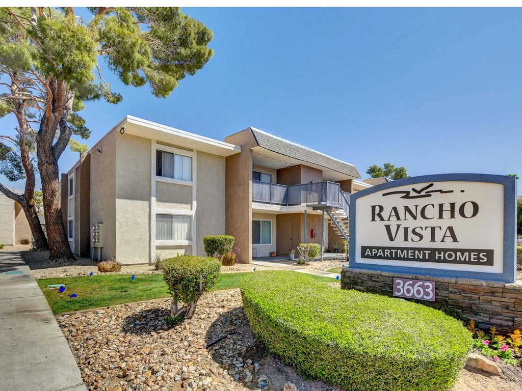 A Rancho Vista Apartment Homes sign is in front of a tan building with a tree and bushes in the front yard.