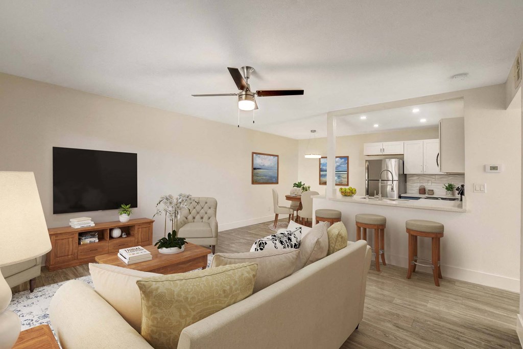 A living room with a beige couch, a wooden coffee table, a flat screen TV, and a kitchen area in the background.