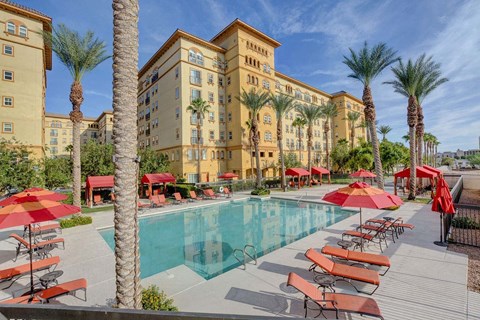 A pool area with red umbrellas and chairs in front of a tan building.