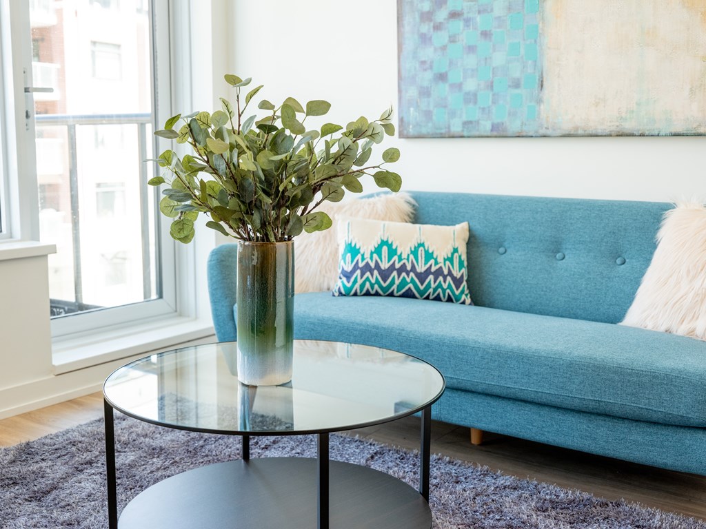 A vase with green plants sits on a glass table in front of a blue couch.