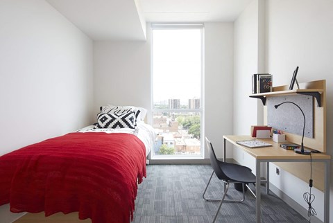 A bedroom with a red bedspread, a desk with a chair, and a window overlooking a cityscape.