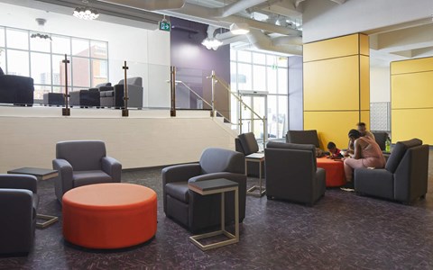 A woman is sitting on a grey chair in a brightly lit room with a purple carpet.